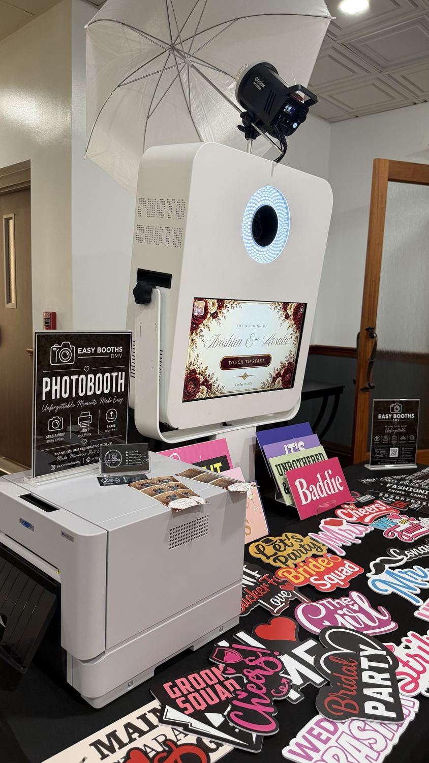 White photo booth with a ring-lit camera, a welcome screen showing a couple's names, a studio flash with umbrella, a printer and a table of props