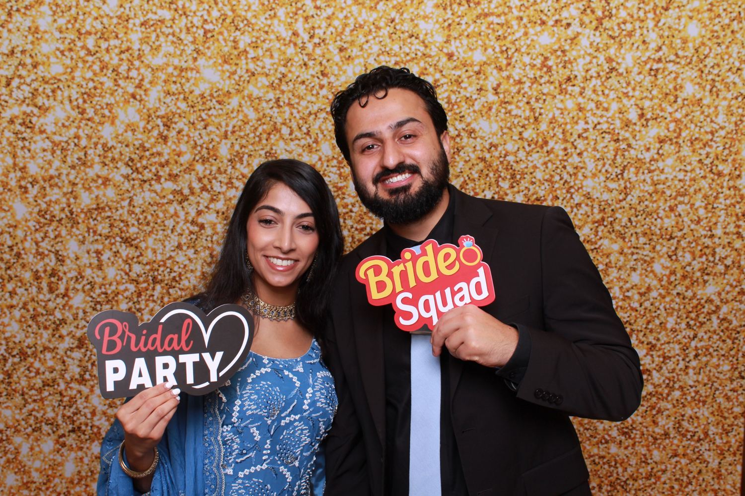Two guests holding Bridal Party and Bride Squad props in front of a gold glitter backdrop