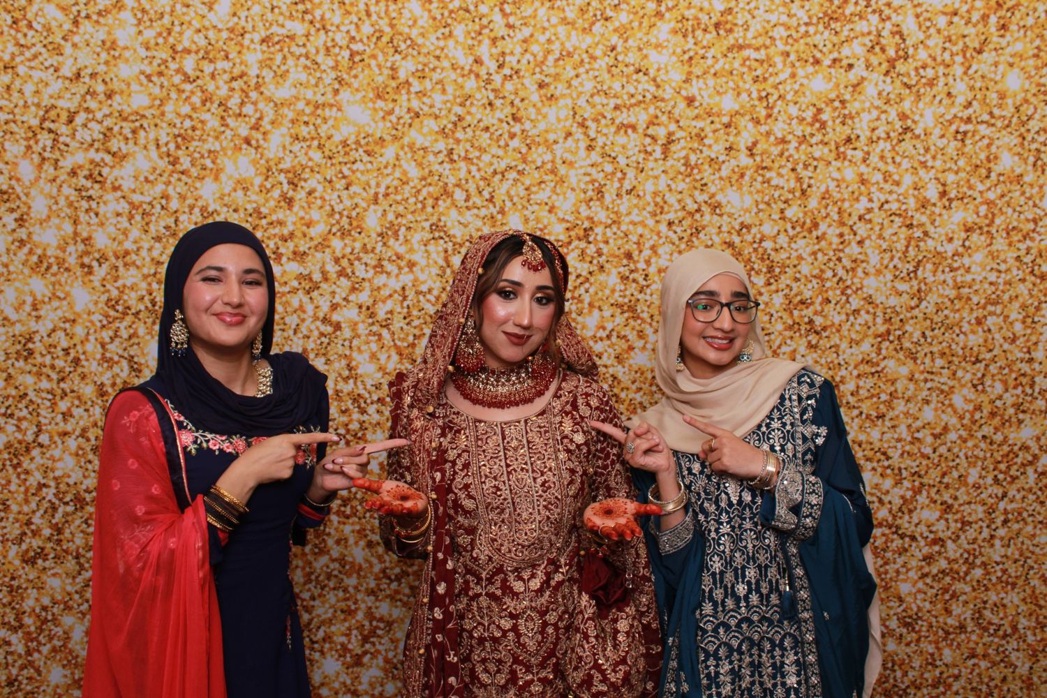 A bride in red and gold with two friends pointing at her henna hands, in front of a gold glitter backdrop