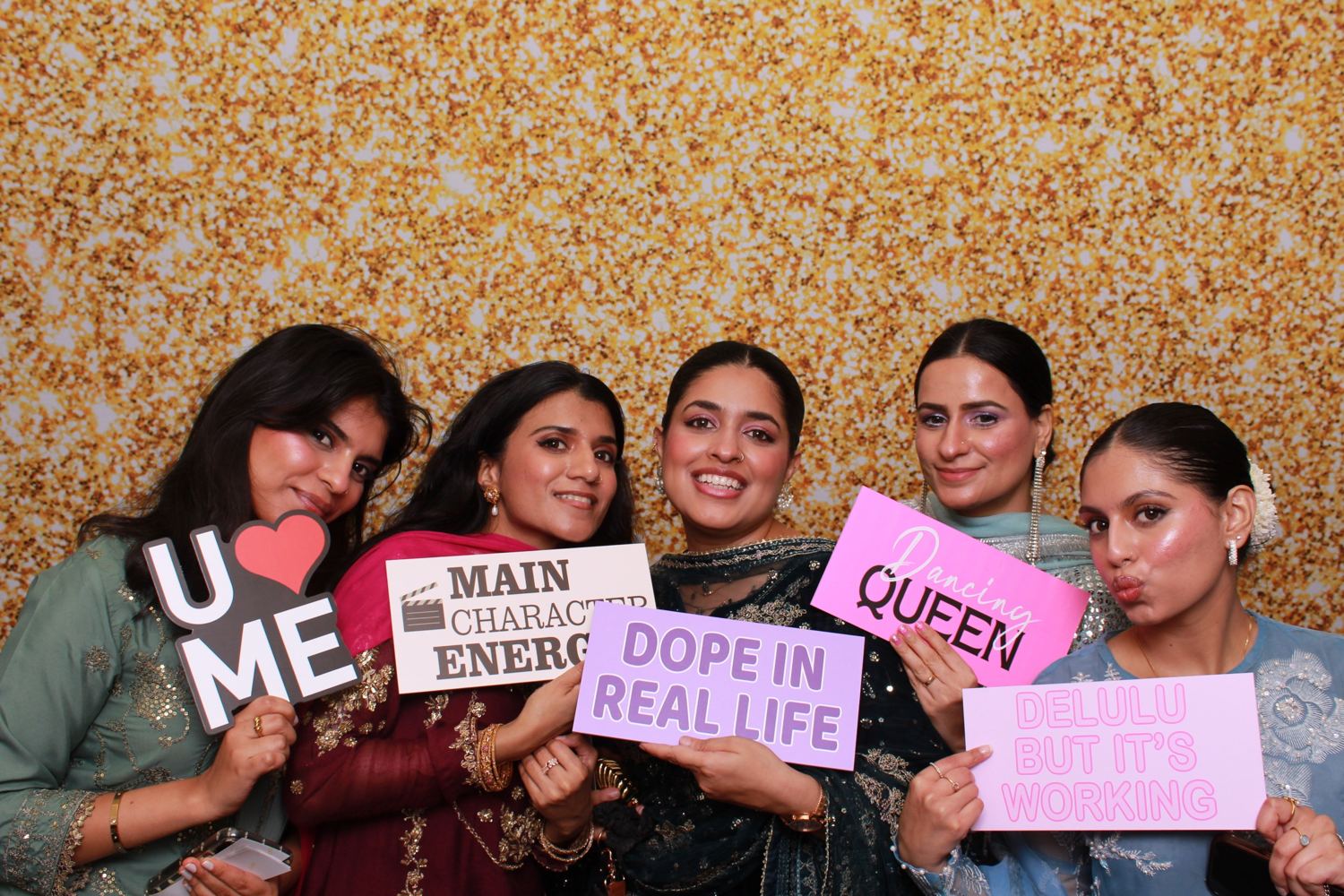Five women in festive outfits holding signs in front of a gold glitter backdrop