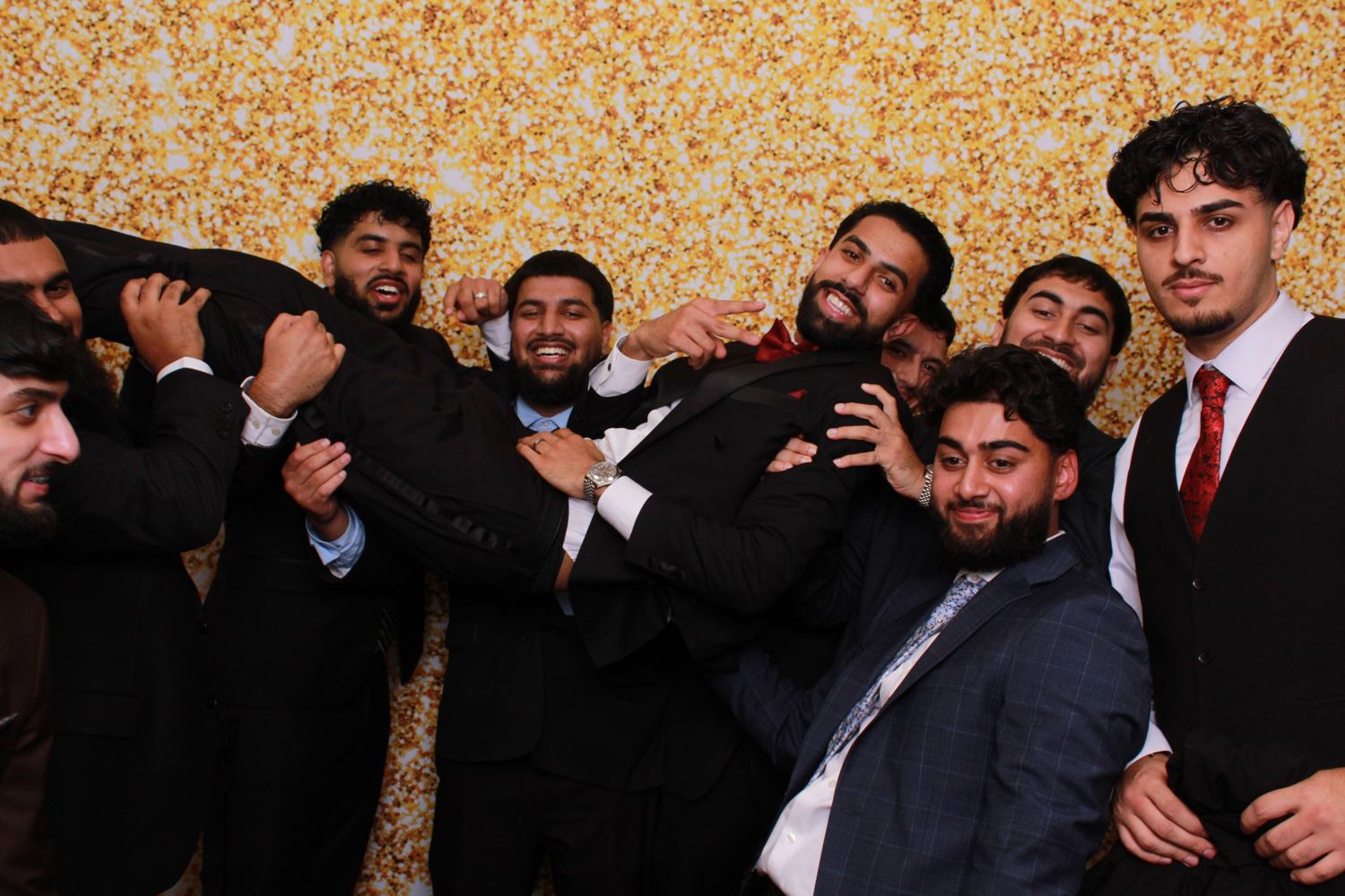 A group of men in suits lifting one of them in front of a gold glitter backdrop