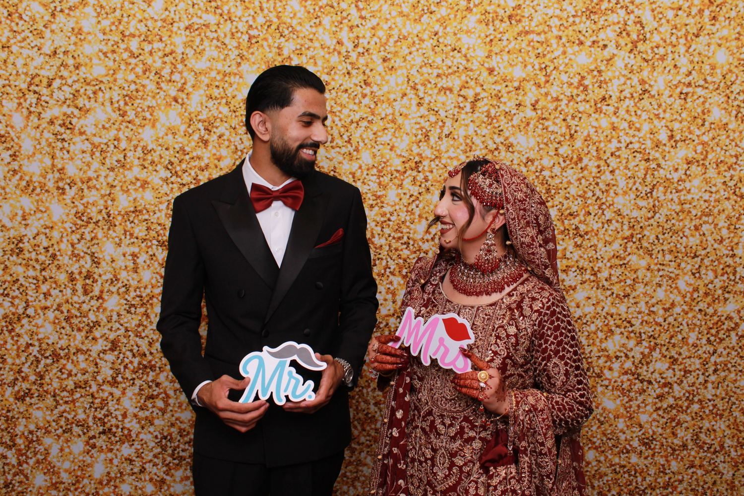A bride and groom holding Mr. and Mrs. props in front of a gold glitter backdrop