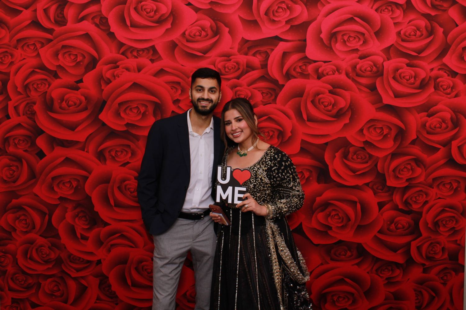 A couple posing in front of a red roses backdrop, holding a U love ME prop