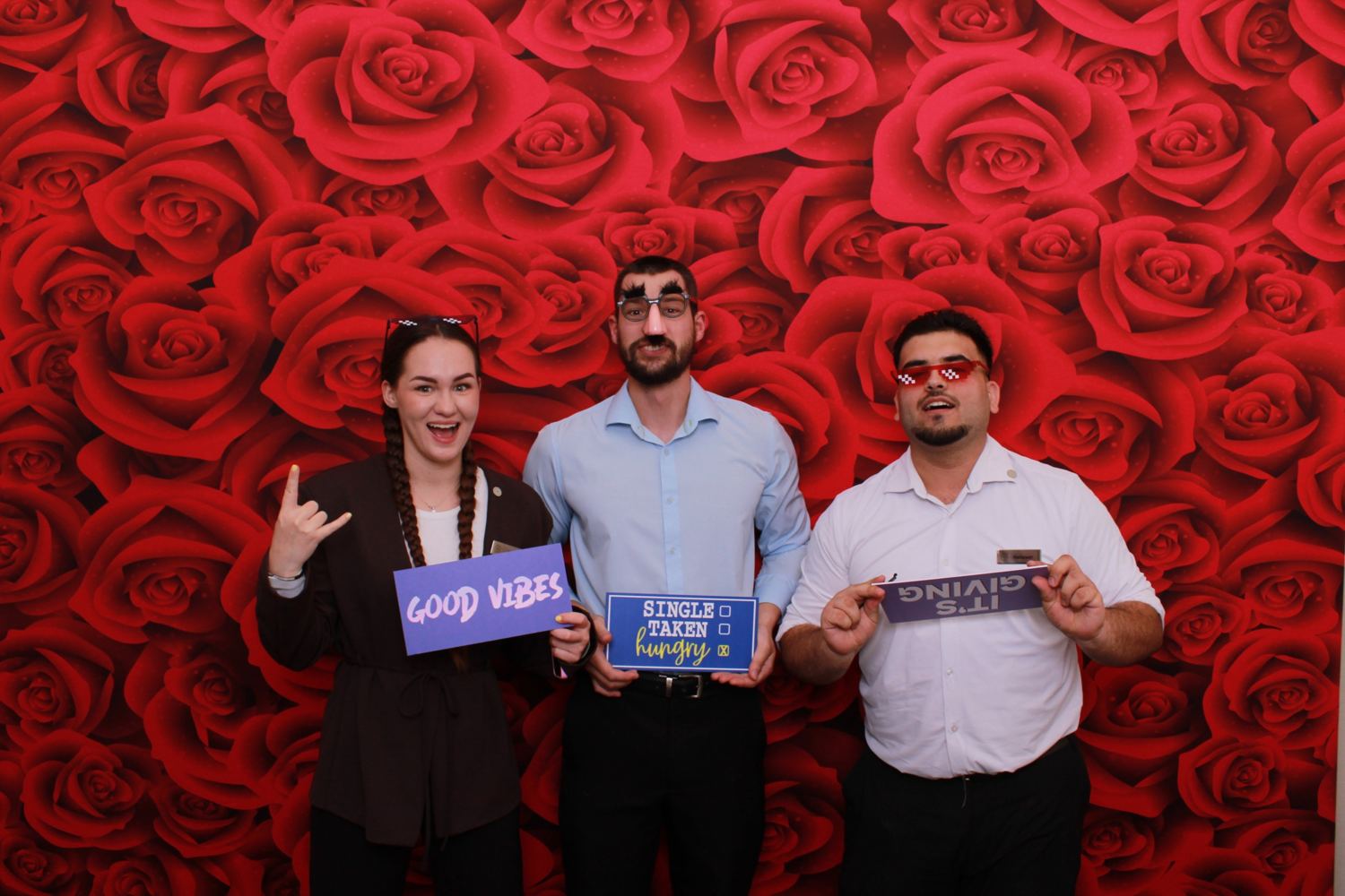 Three coworkers in novelty glasses holding signs in front of a red roses backdrop