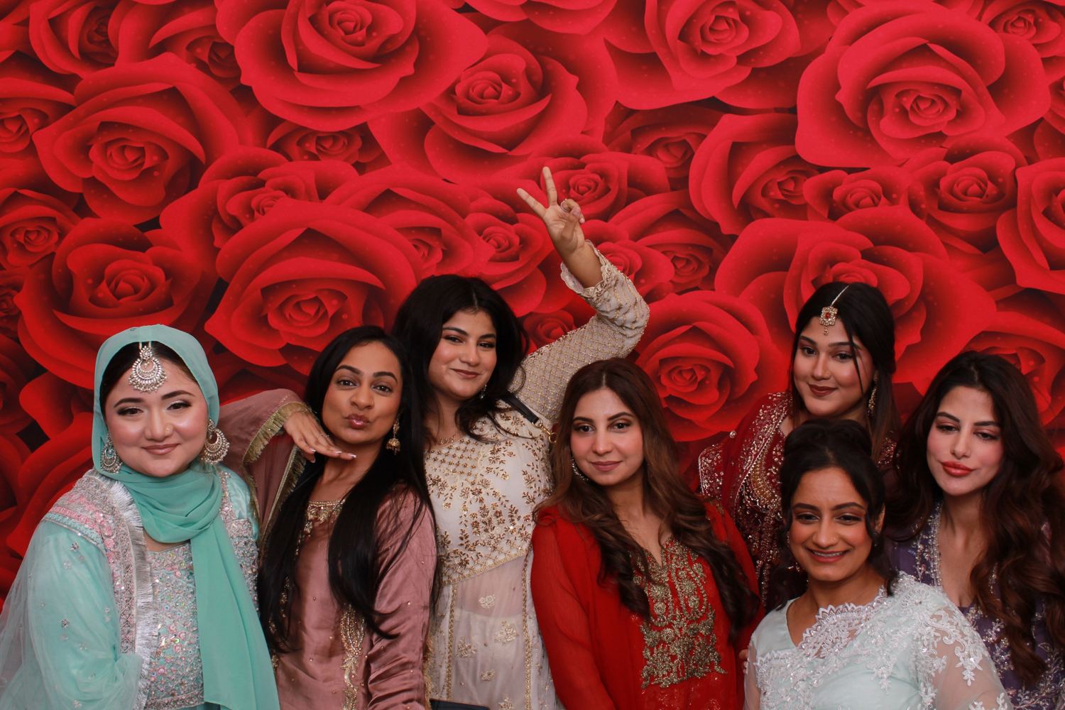 Seven women in embroidered outfits posing in front of a red roses backdrop