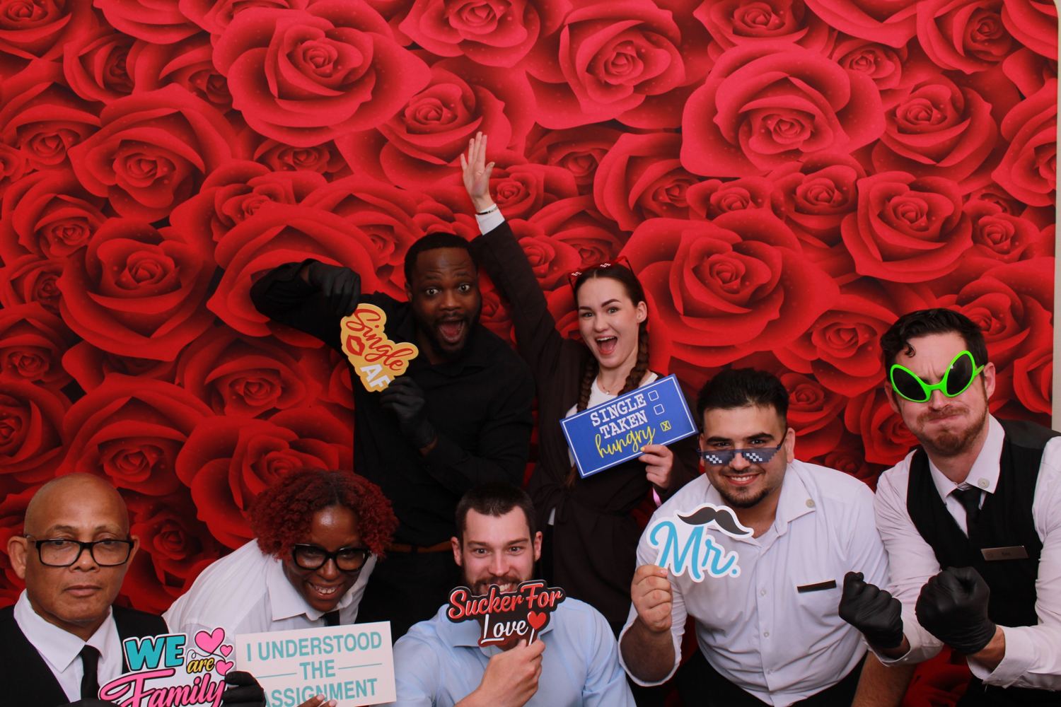 Seven people in work uniforms striking silly poses with props in front of a red roses backdrop