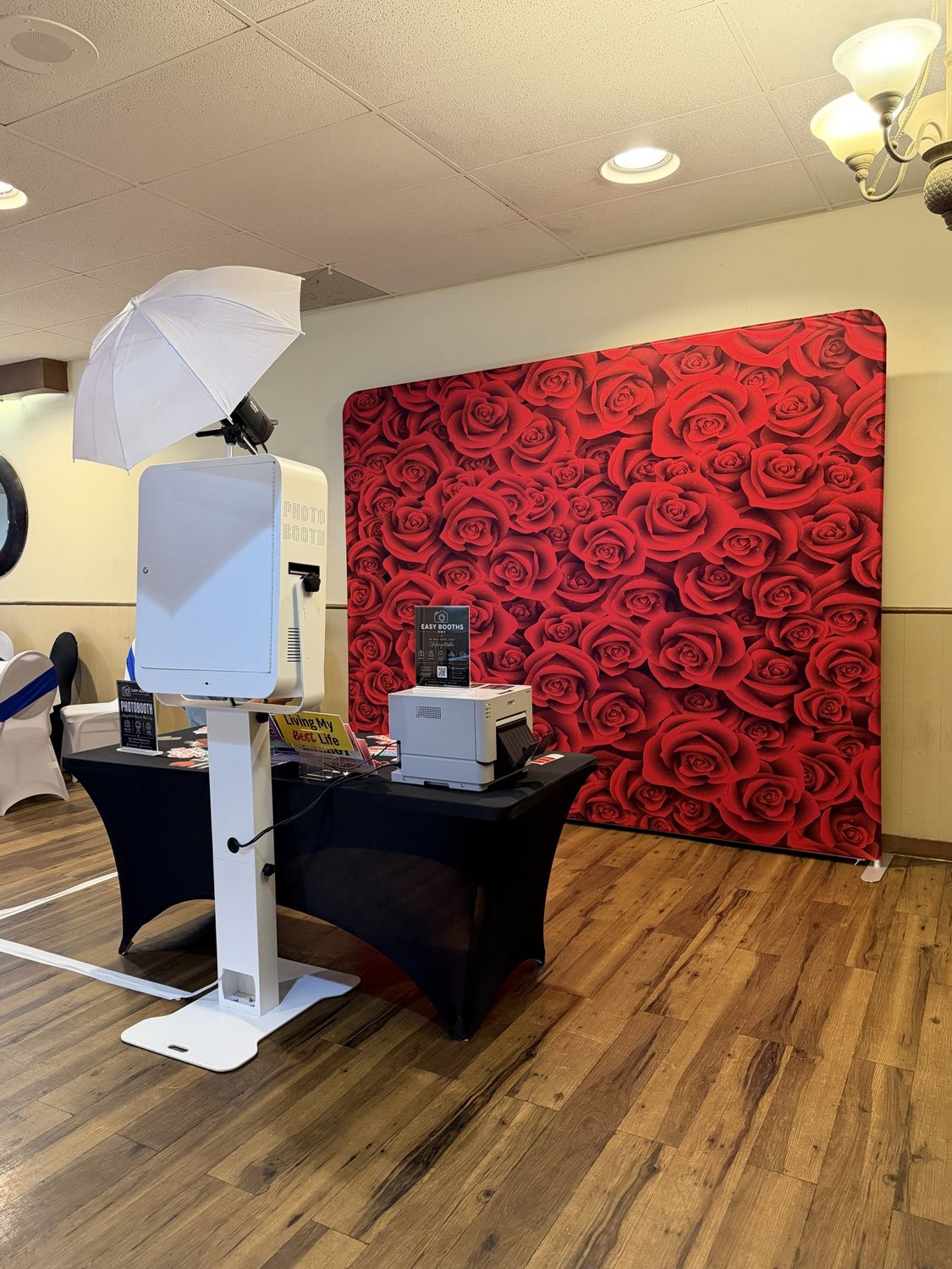 Photo booth, printer and prop table beside a red roses backdrop on a wood floor
