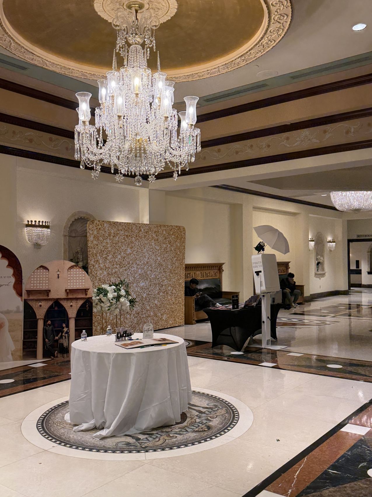 Photo booth, printer and a gold glitter backdrop set up in a hall lobby under a chandelier, with open floor around them
