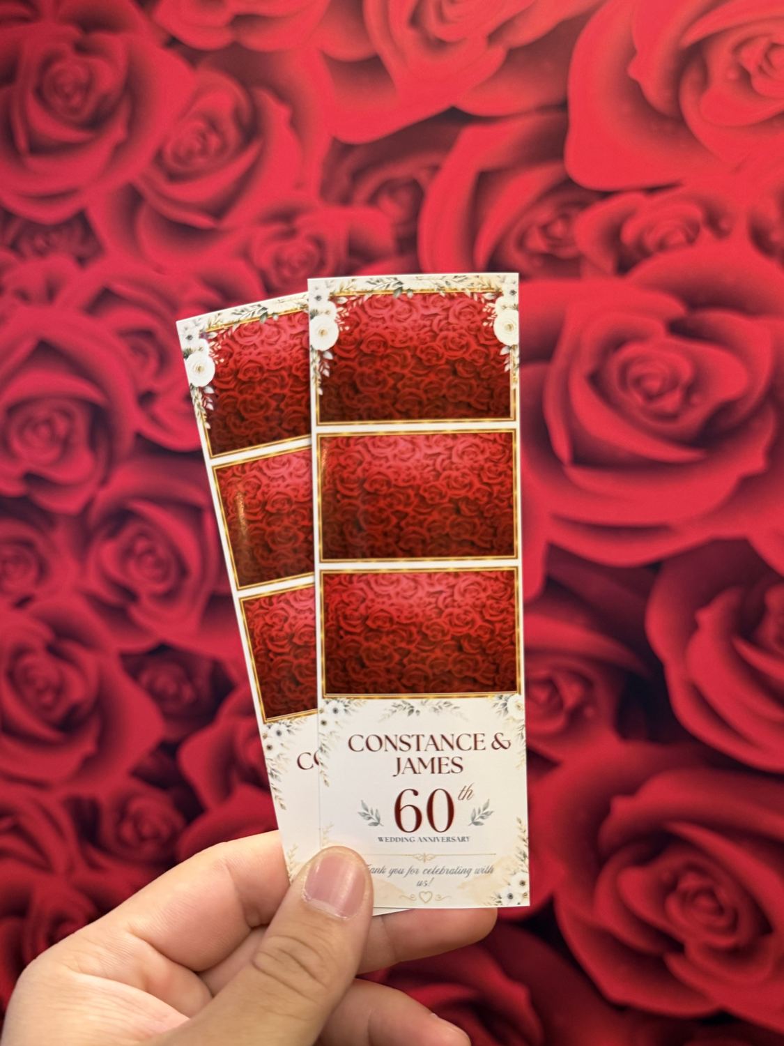 A hand holding two 60th wedding anniversary photo strips with white flowers, in front of a red roses backdrop